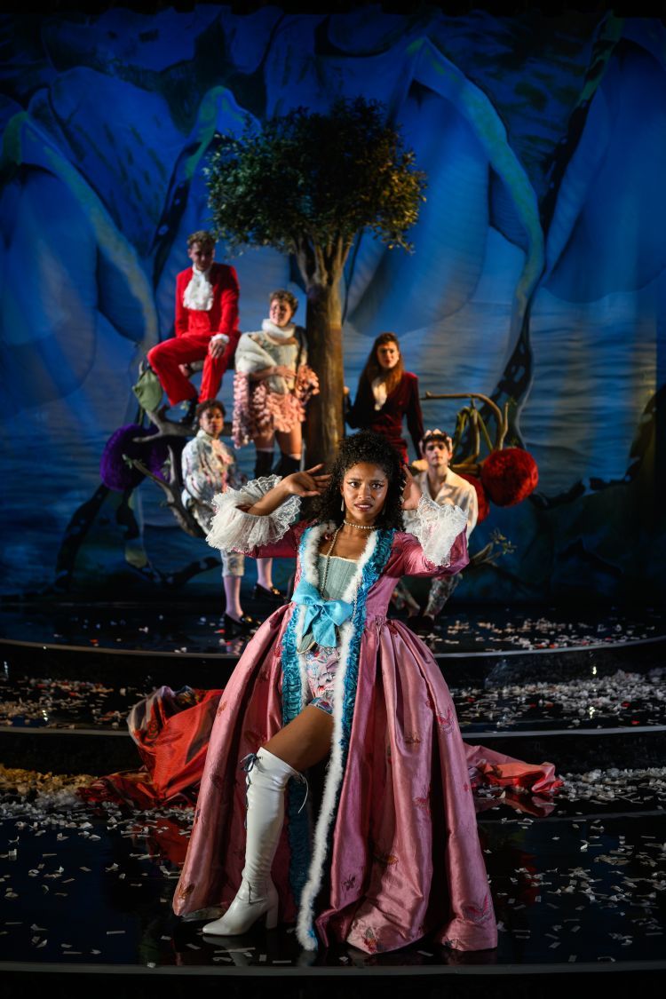 Actor playing Orlando is walking across the front of the stage with a silk pastel pink renaissance style gown with a white fluffy trim. Other cast member sit on the branches of a tree in the background in red 1970's style suits with white shirts/ruffles or blue and pink pastel costumes to represent Orlando.
